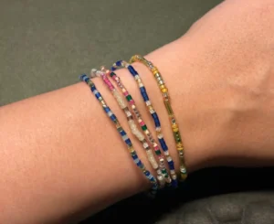 Close-up of several handmade beaded bracelets in blue, pink, white, green and gold tones worn on a wrist.